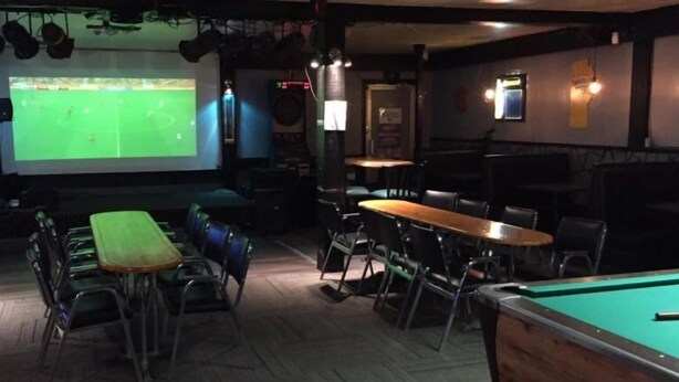 Uma sala de bilhar com mesa de sinuca, tela de projeção exibindo um jogo de futebol e algumas mesas e cadeiras.