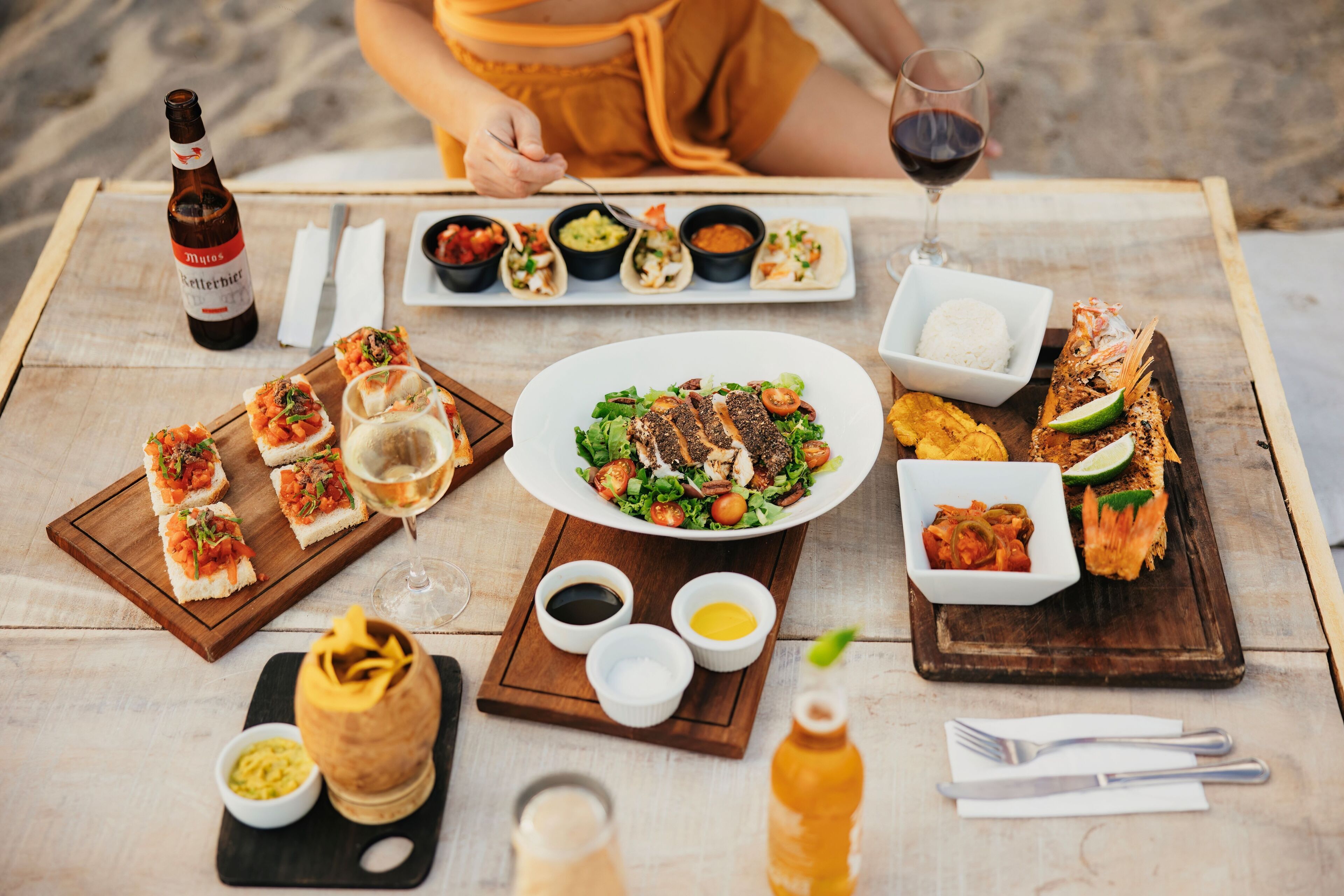 A wooden table with various dishes, including tacos, a salad, and grilled fish, along with condiments and drinks.