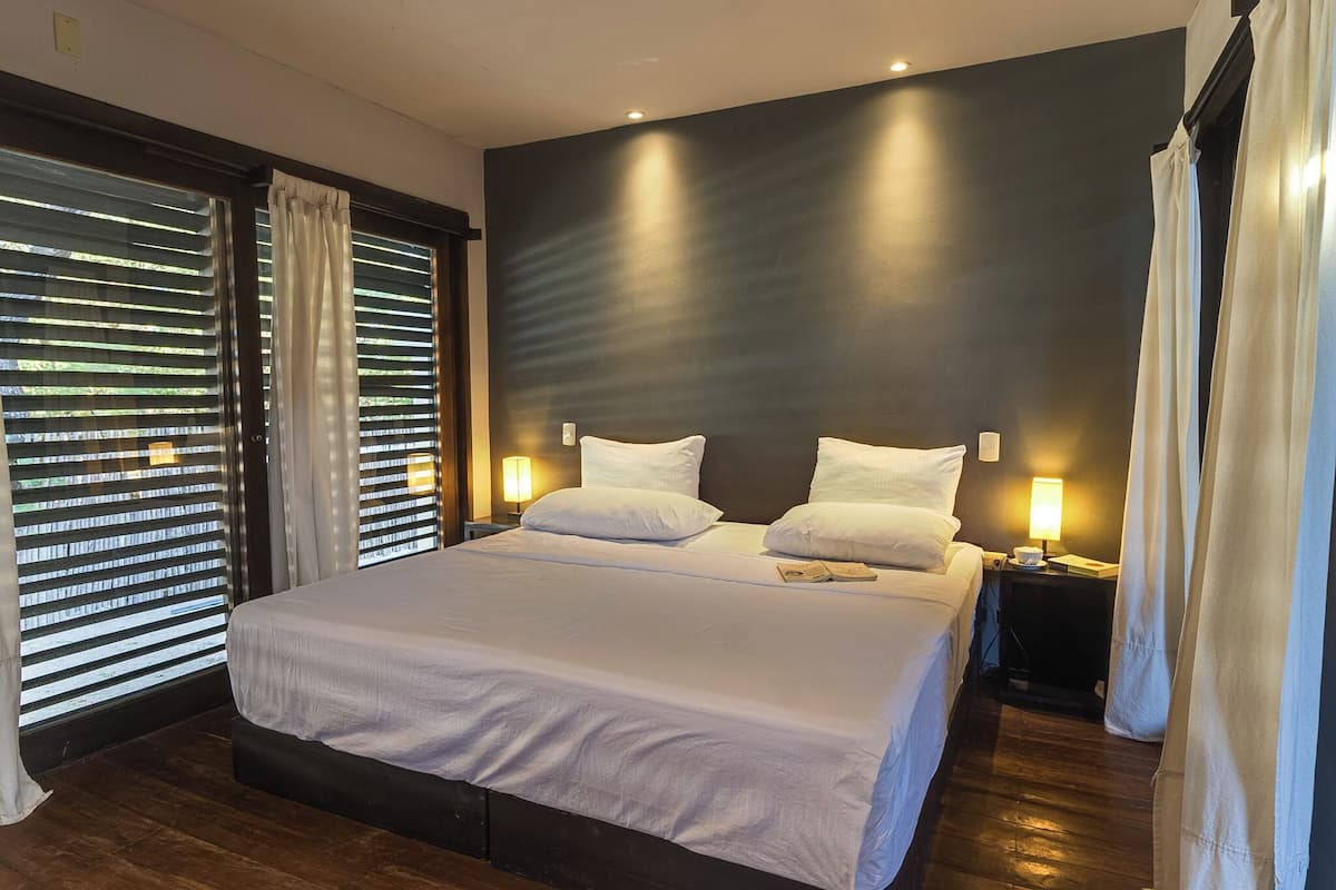 A modern hotel room with a large bed, bedside lamps, and a view through the window blinds.