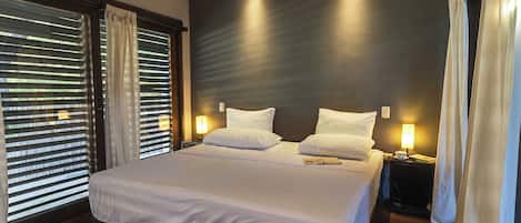 A modern hotel room with a large bed, bedside lamps, and a view through the window blinds.