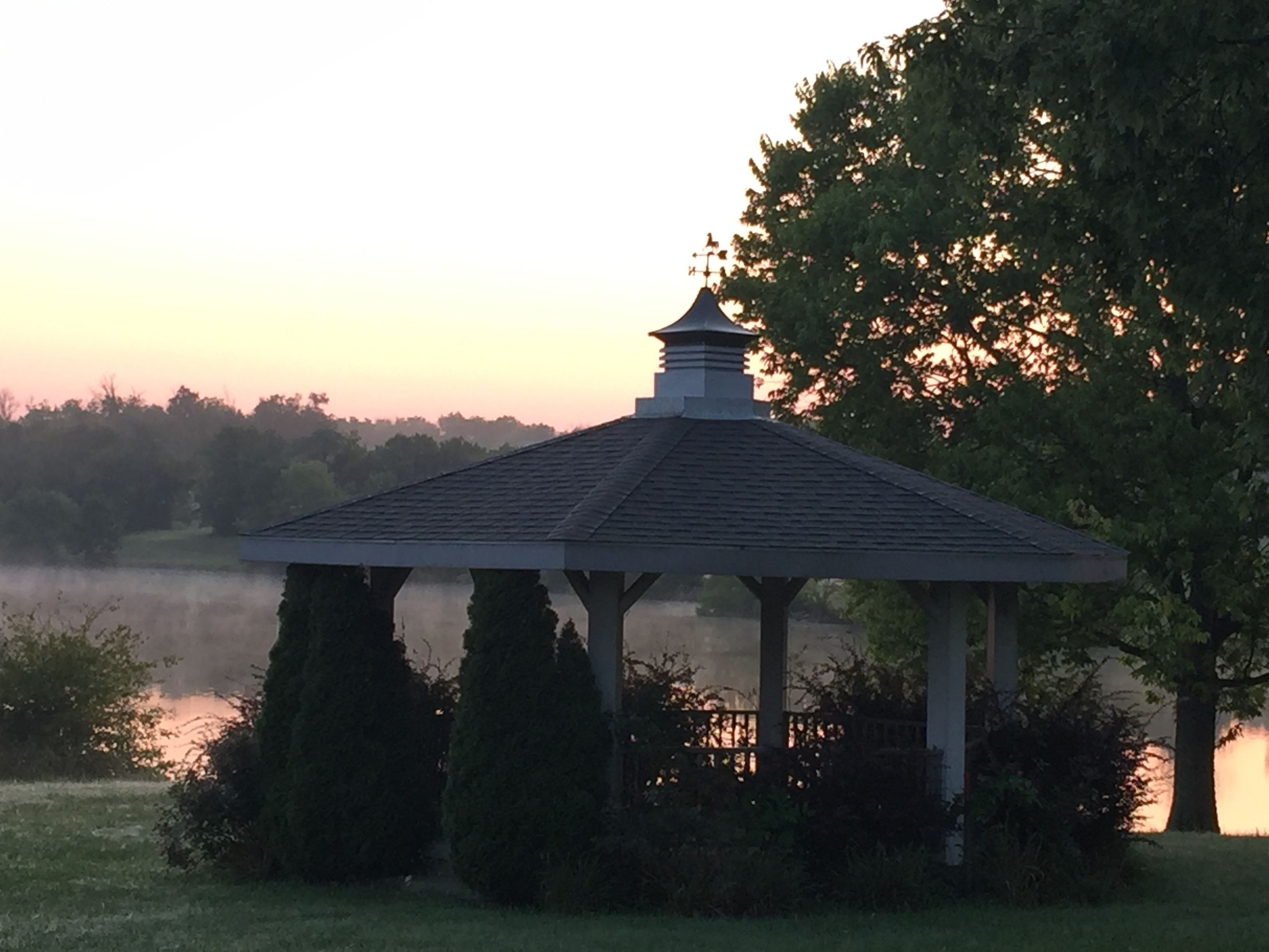 A gazebo with a tiled roof, surrounded by trees and a body of water.