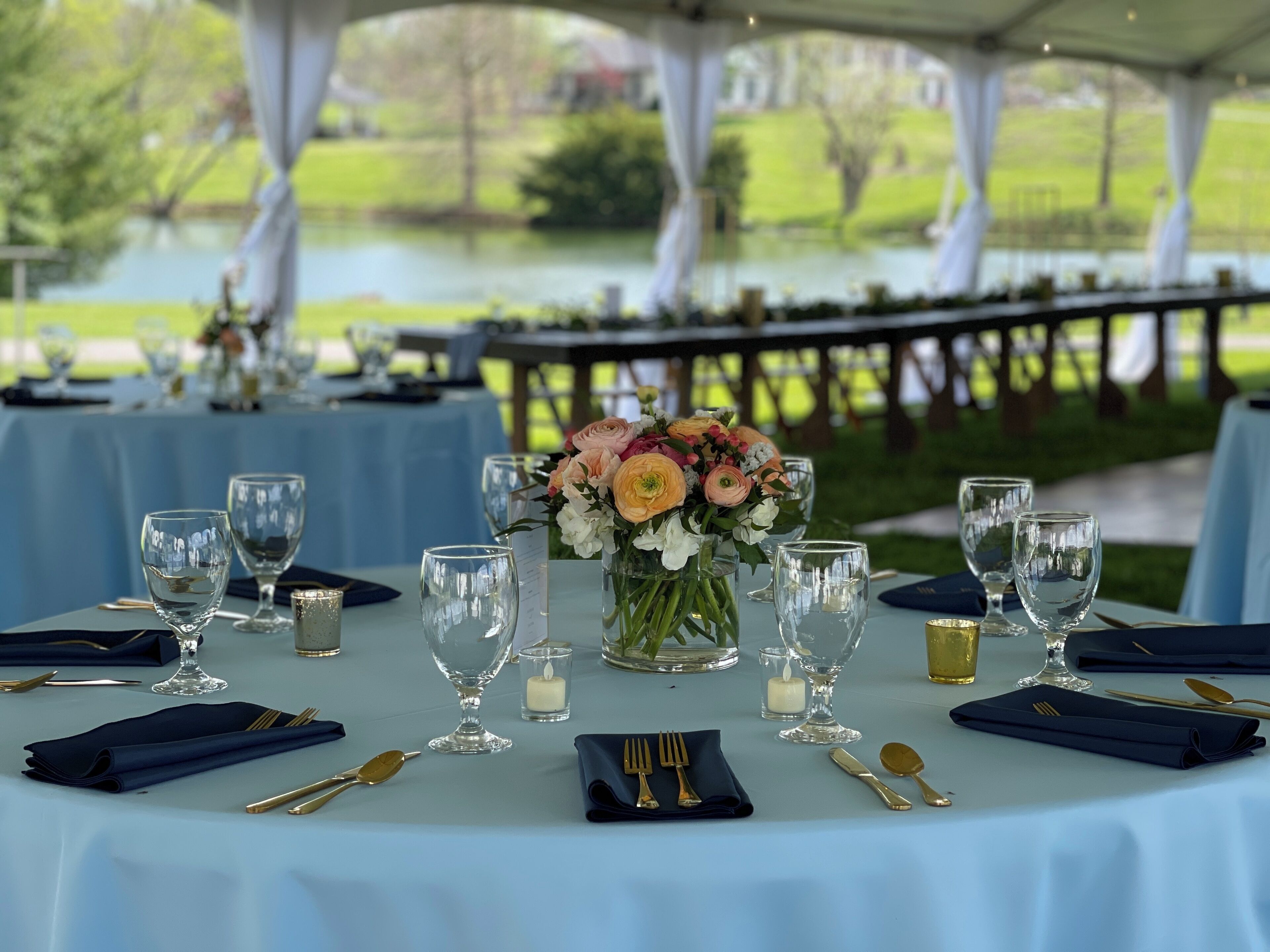 Mesa posta para um evento ao ar livre formal, com vista para um lago, exibindo toalha azul, copos, talheres e um arranjo de flores.