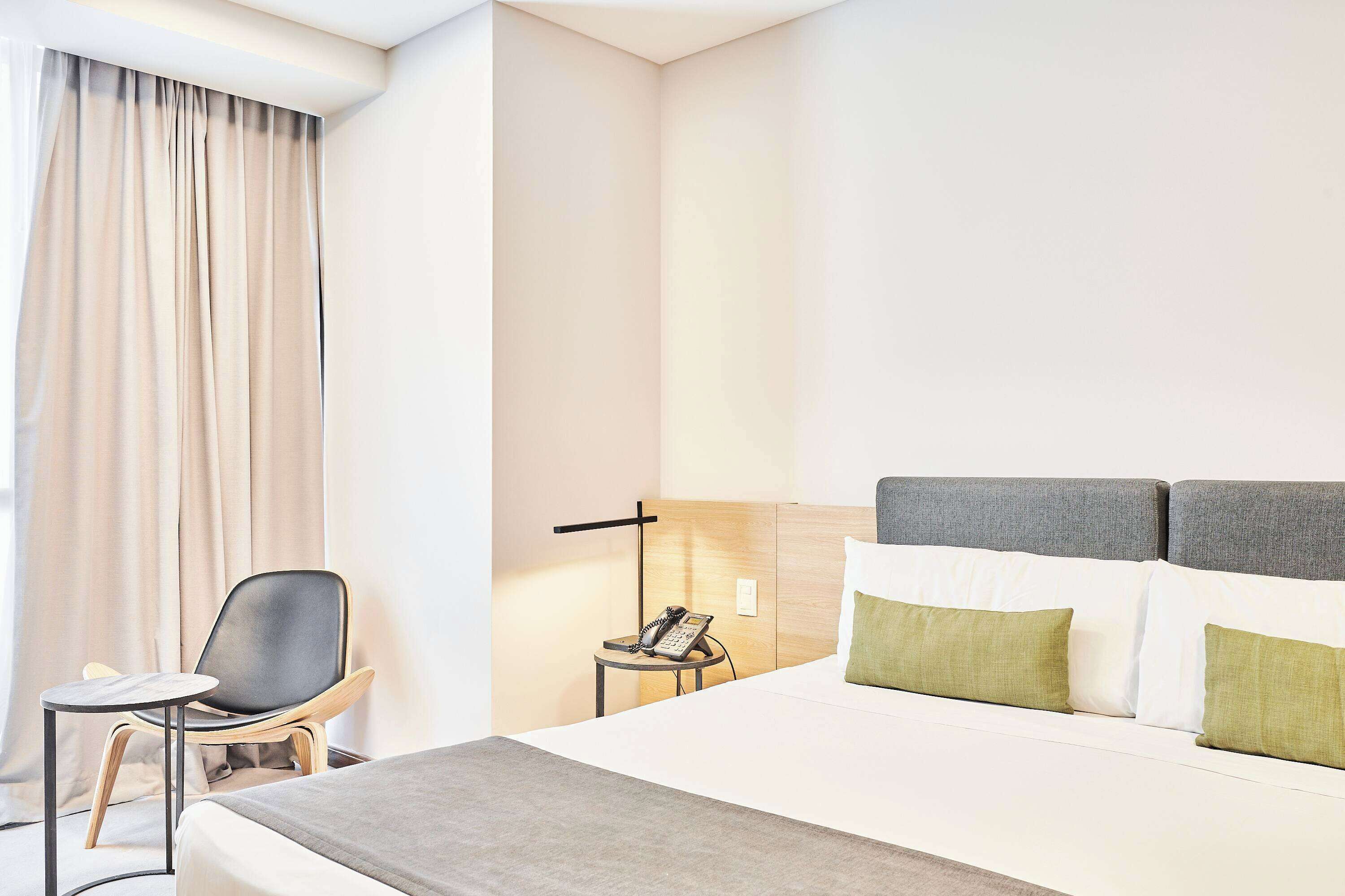 A modern hotel room with a bed, a chair, a bedside table with a phone, and a lamp.