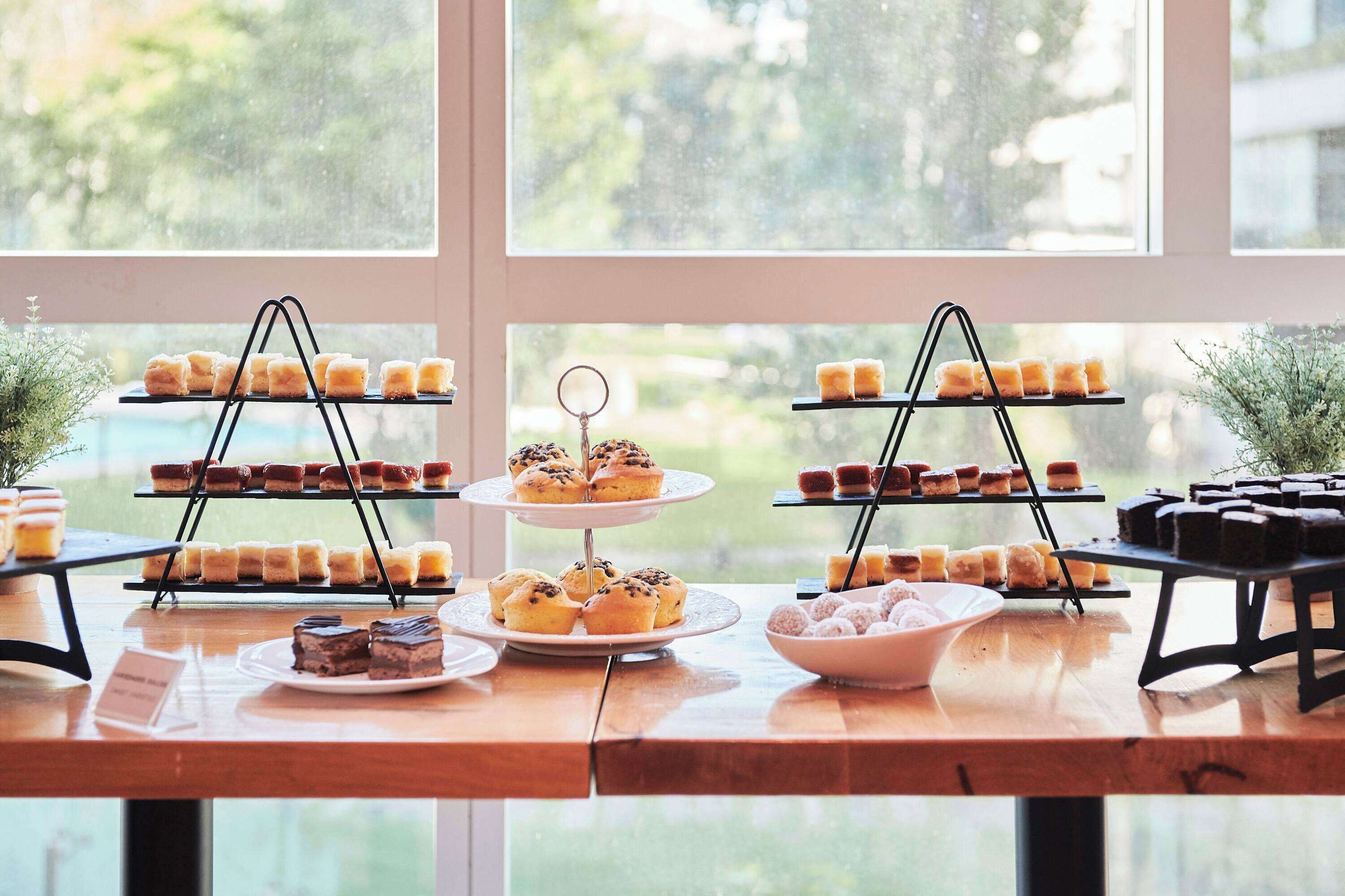 Una mesa de madera exhibe varios niveles de pasteles y tartas, cerca de amplios ventanales.