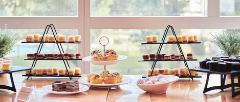 Una mesa de madera exhibe varios niveles de pasteles y tartas, cerca de amplios ventanales.