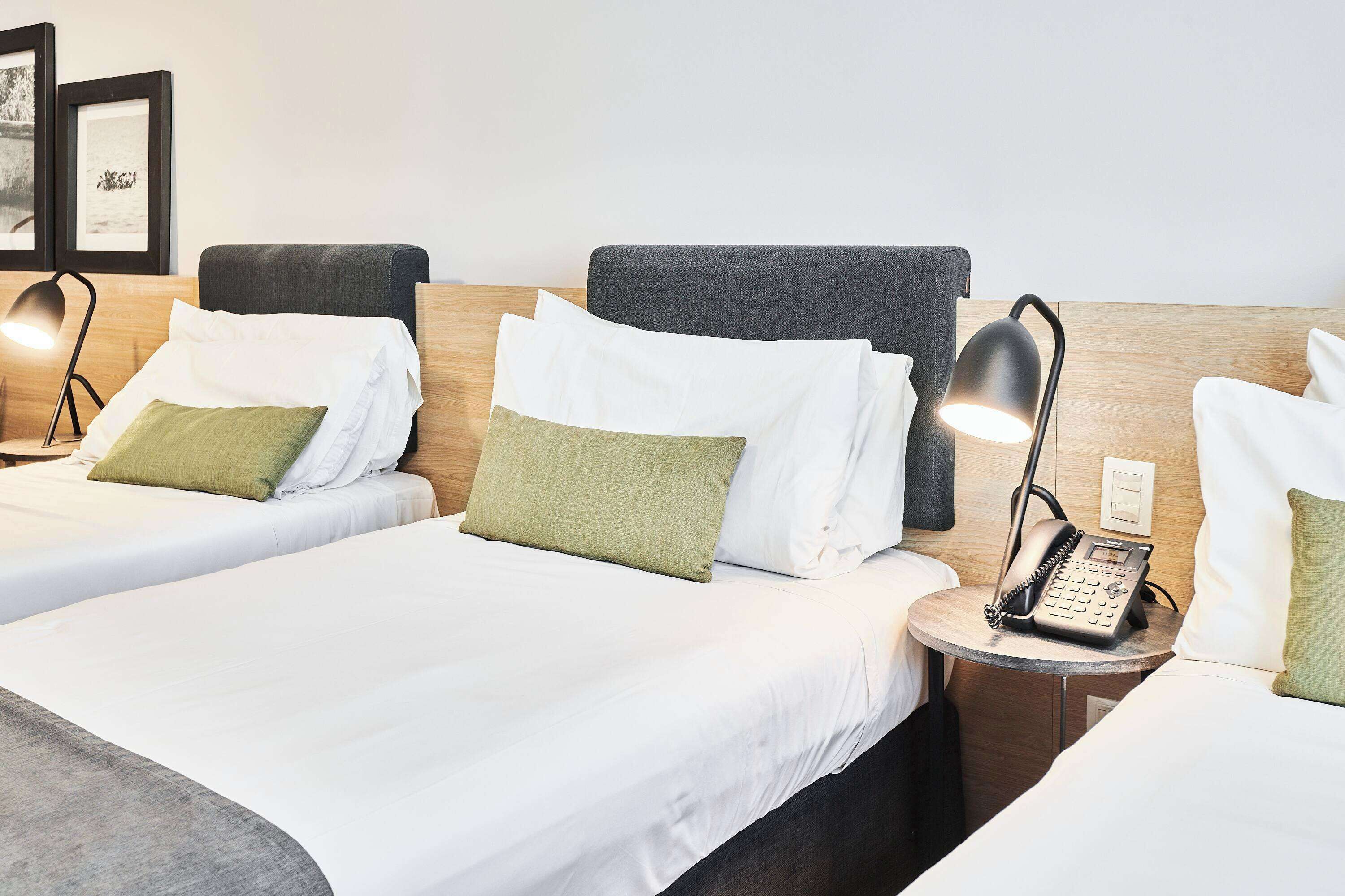 A hotel room with two beds, each with white linens and green pillows, a bedside table with a lamp and a telephone, and framed pictures on the wall.