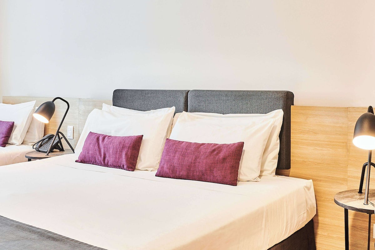 Ein ordentlich gemachtes Bett mit weißen und violetten Kissen, einem grauen Kopfteil und einer Nachttischlampe.