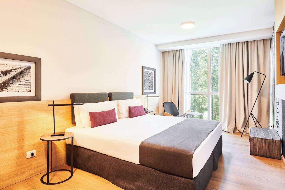Ein Hotelzimmer mit einem großen Bett, einem Nachttisch, einem Sessel, einem Schreibtisch, einer Lampe und einem Fenster mit Vorhängen.