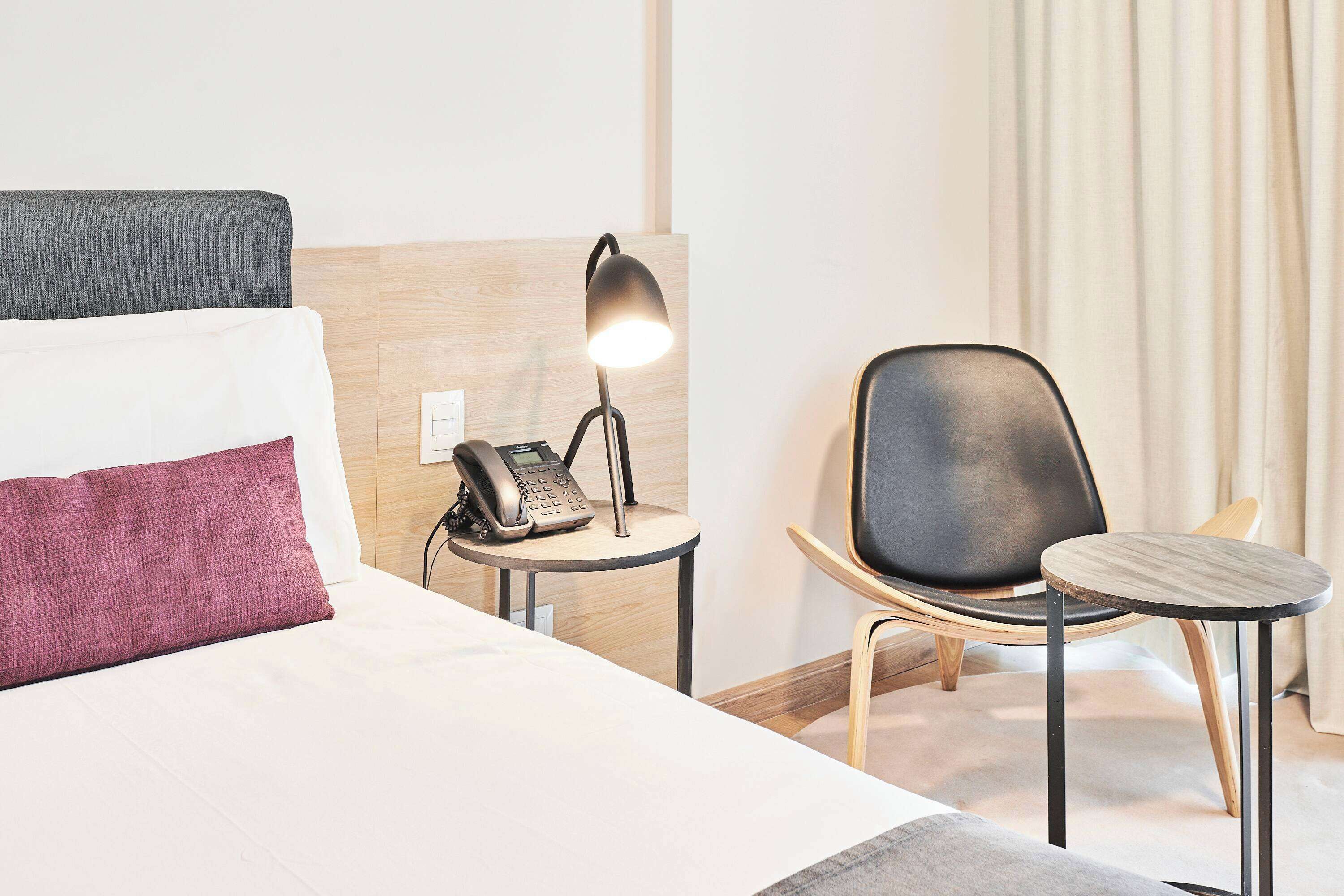 Ein Hotelzimmer mit Bett, Nachttischtelefon, Lampe, Stuhl und kleinem Tisch.
