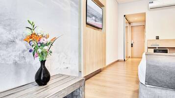 A hotel hallway with a wooden bench, a vase of flowers, a framed picture on the wall, and a bed with a grey bedspread.