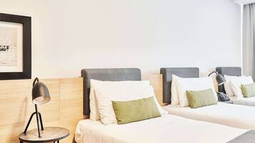 Ein Hotelzimmer mit zwei Betten, jeweils mit grauem Kopfteil, weißer Bettwäsche und einem grünen Kissen. Neben den Betten steht ein runder Tisch mit einer Lampe.