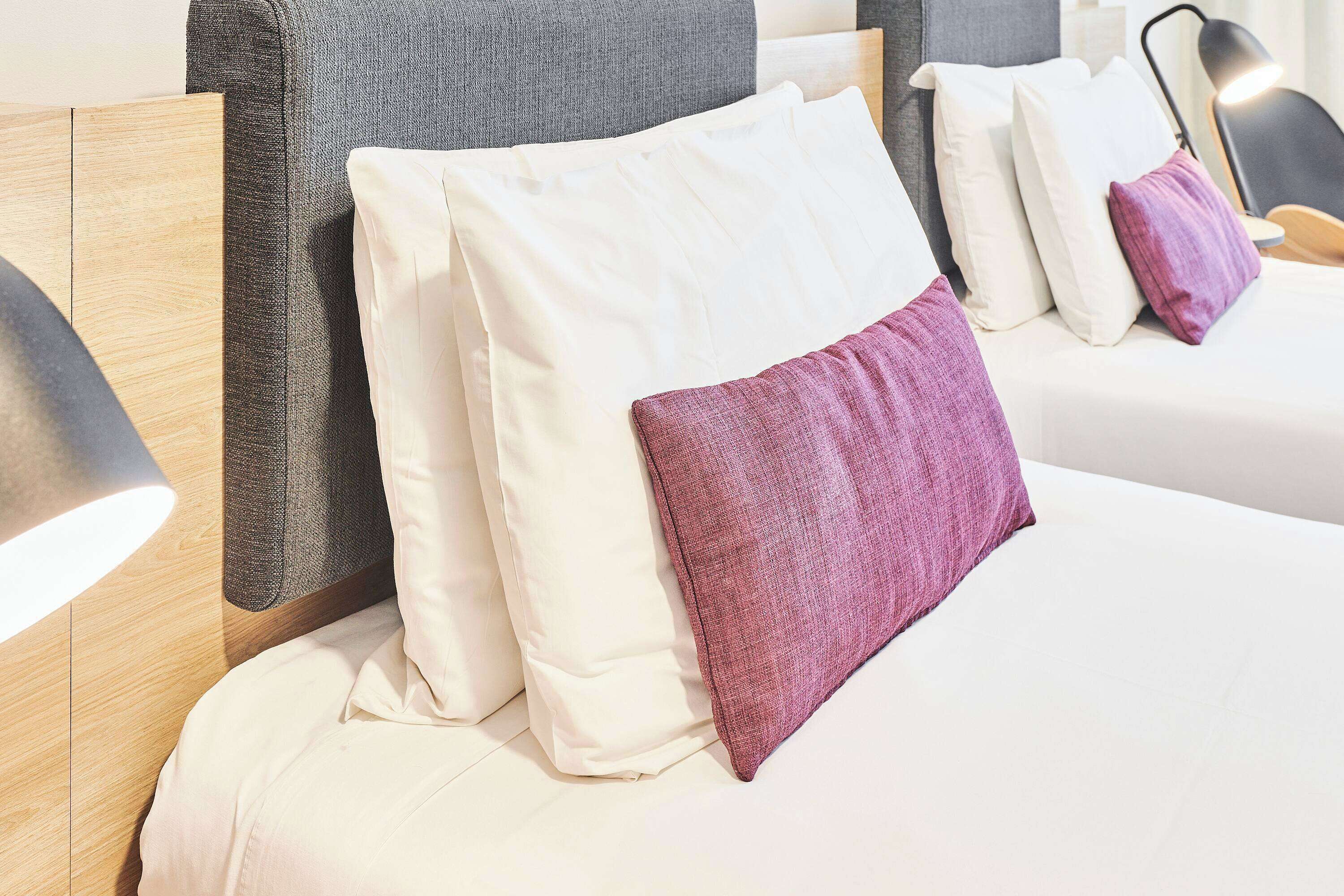 A neatly made bed with white and purple pillows, a wooden headboard, and a bedside lamp.