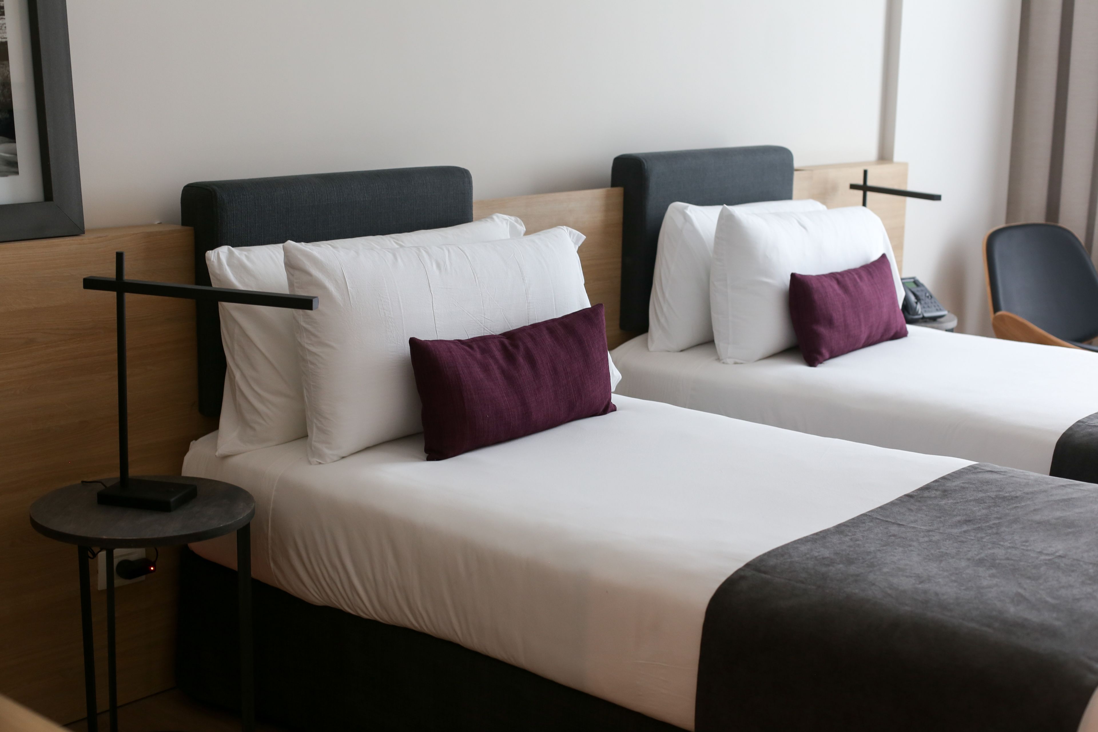 Two beds with white linens and dark headboards, a small round table with a power outlet, and a chair.