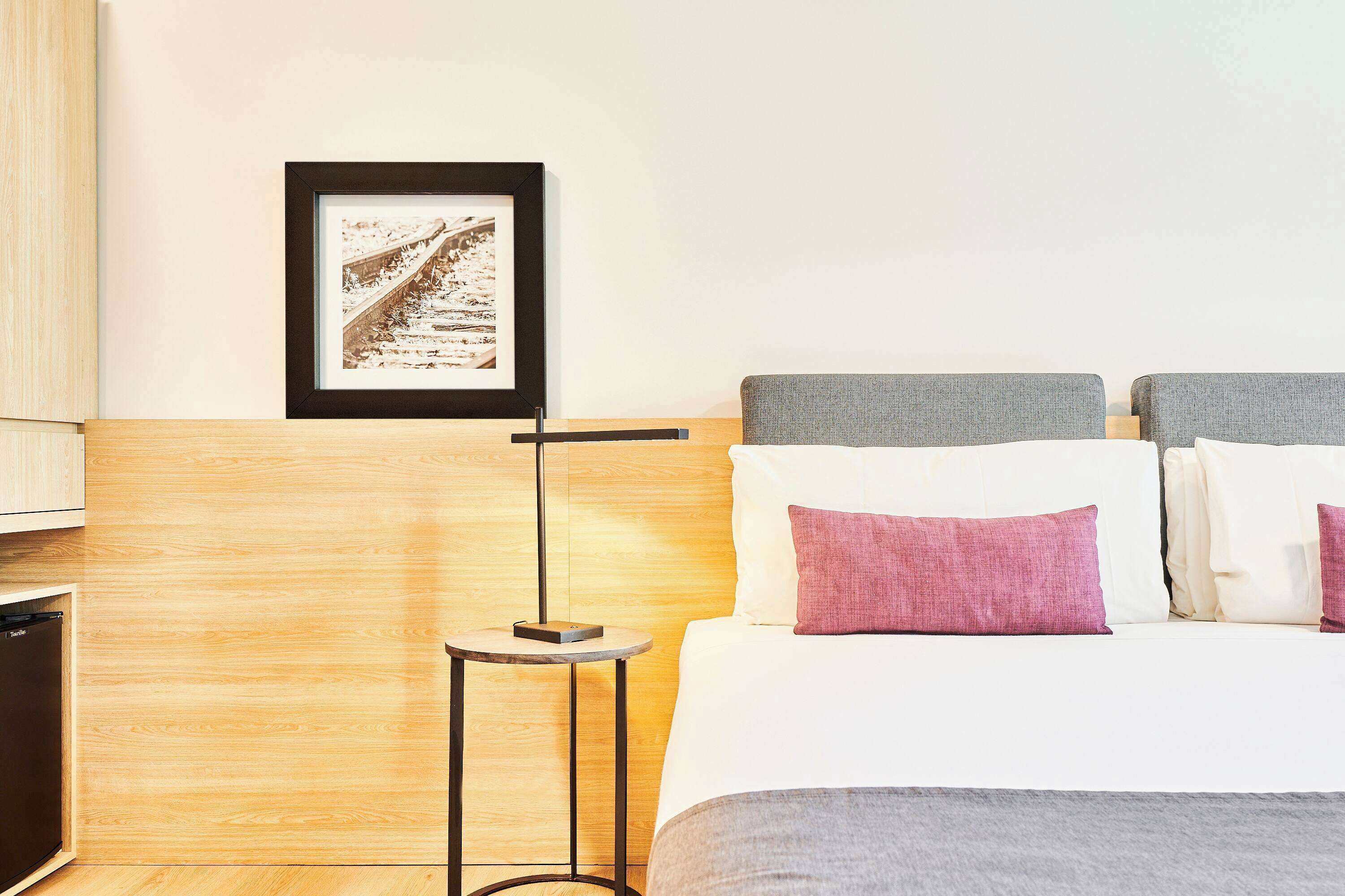 Une chambre d’hôtel bien rangée, avec un lit, des oreillers, une table de chevet, un tableau encadré et une lampe sur pied.
