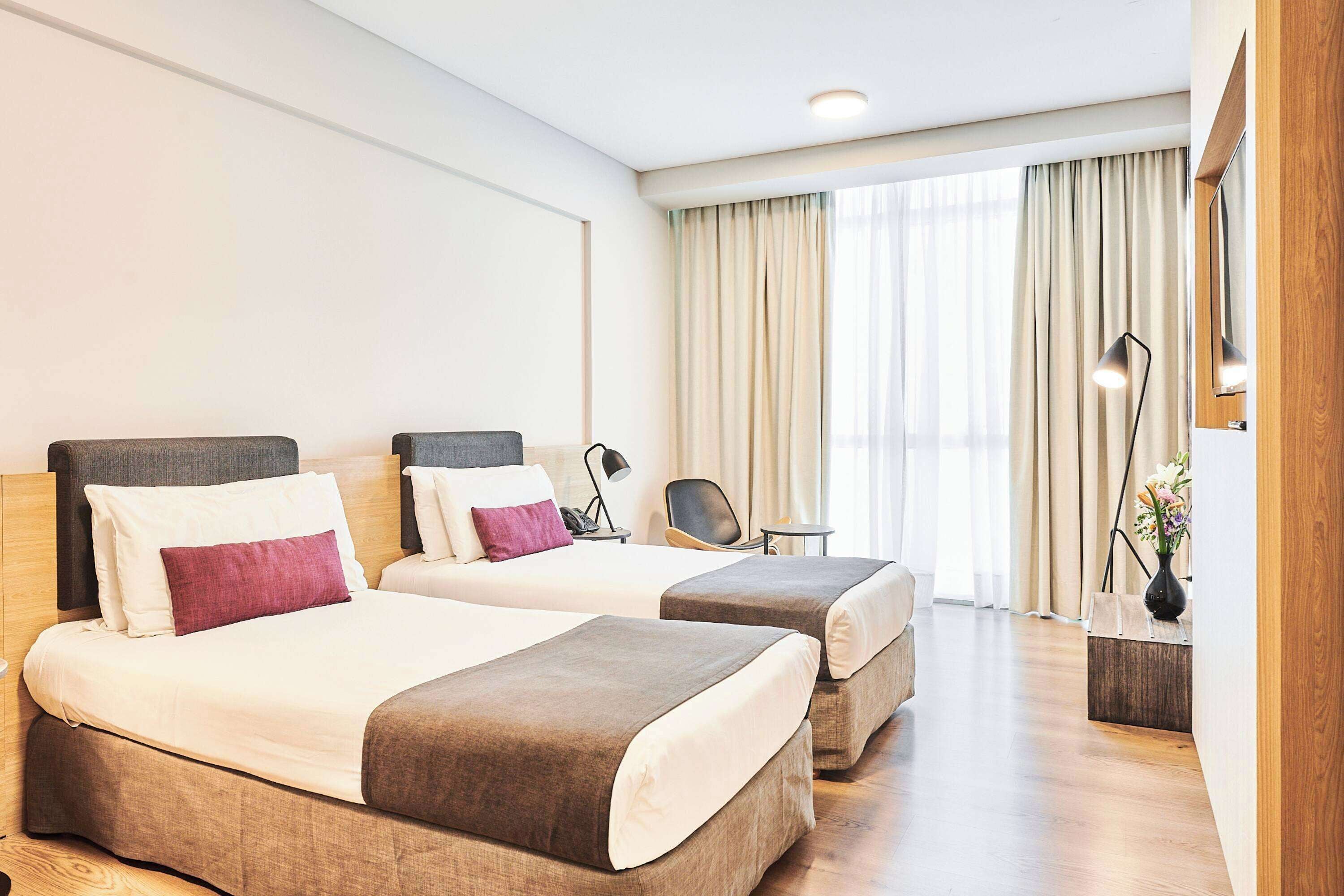 Una habitación de hotel con dos camas, un escritorio, una silla, una lámpara y una ventana con cortinas.
