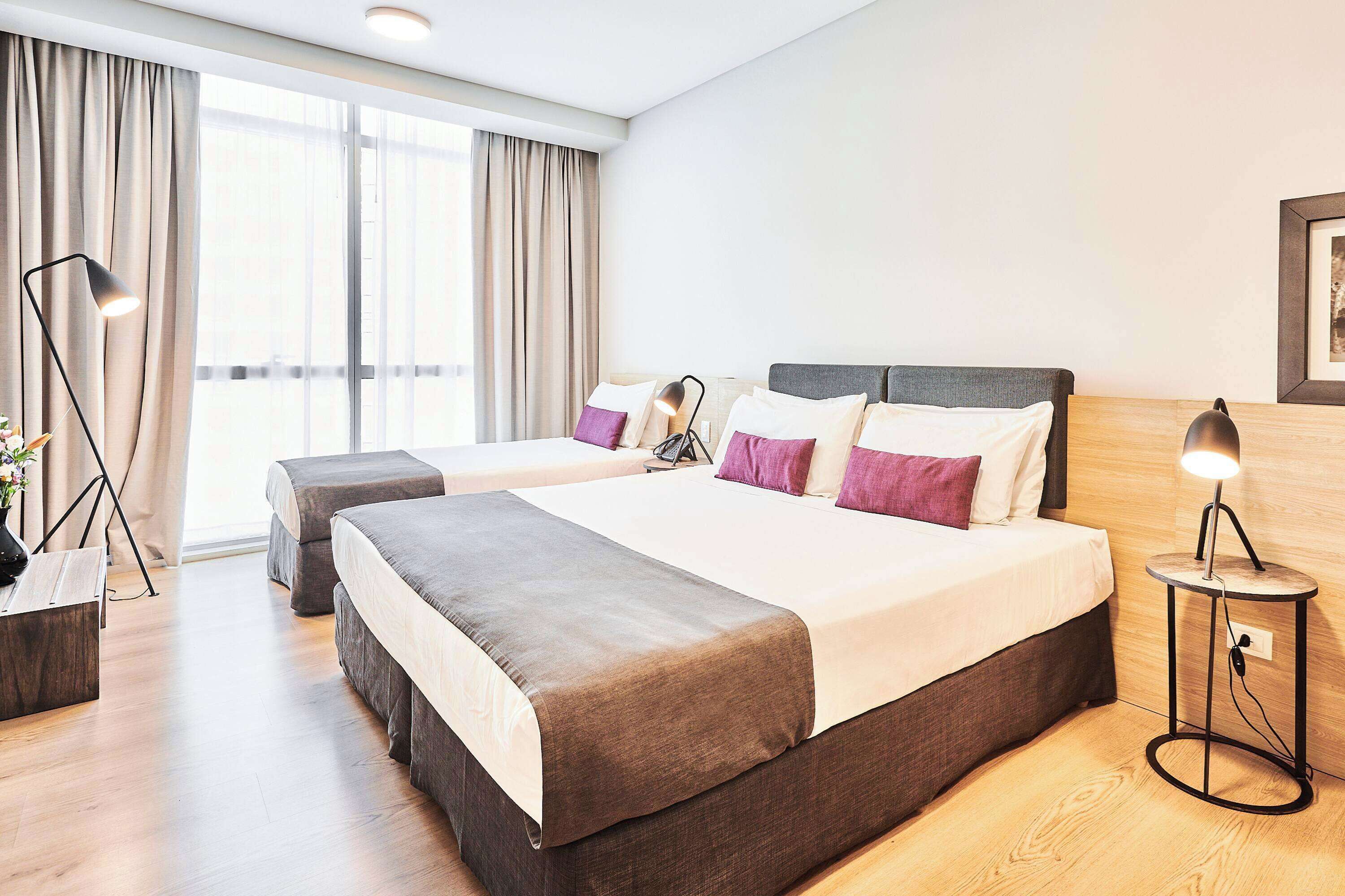 A modern hotel room with two beds, a large window, and a bedside table with a lamp.