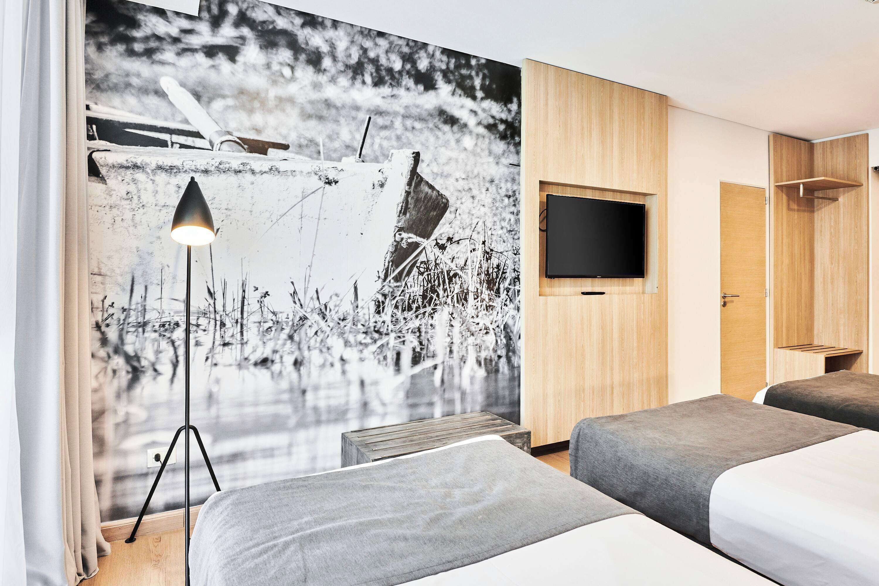 Habitación de hotel con dos camas, un televisor de pared grande y un mural de un barco en el agua.