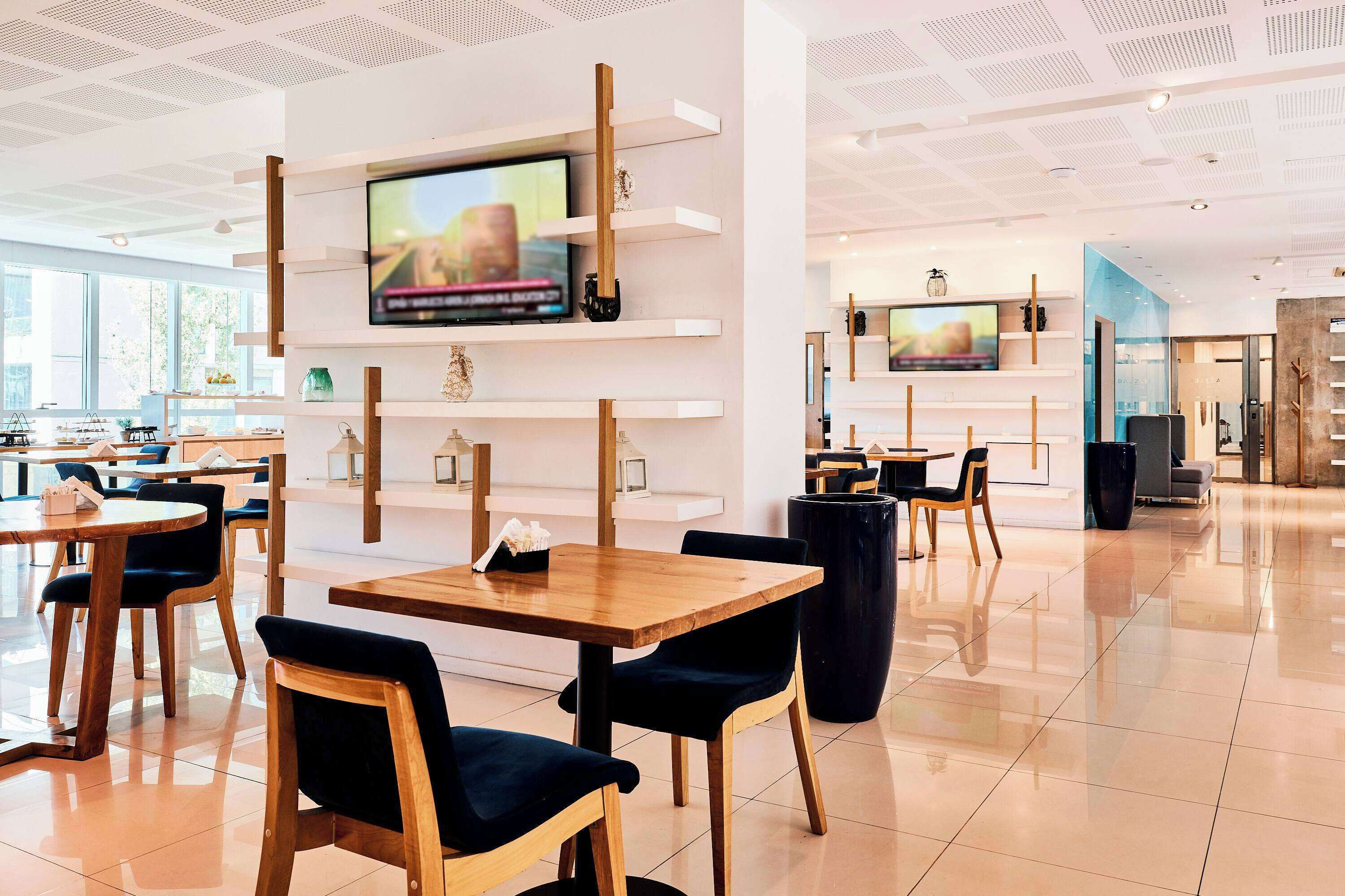 Un restaurant moderne avec des tables et des chaises en bois, un téléviseur fixé à un mur blanc et un intérieur lumineux et spacieux.