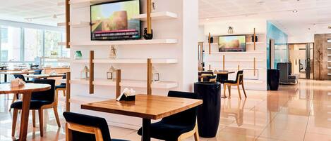 A modern restaurant with wooden tables and chairs, a TV mounted on a white wall, and a bright, spacious interior.