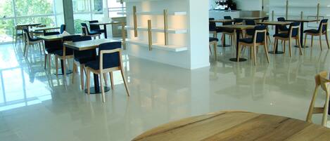 A modern dining area with wooden tables and chairs, a kitchen area with a coffee machine, and a large window providing natural light.