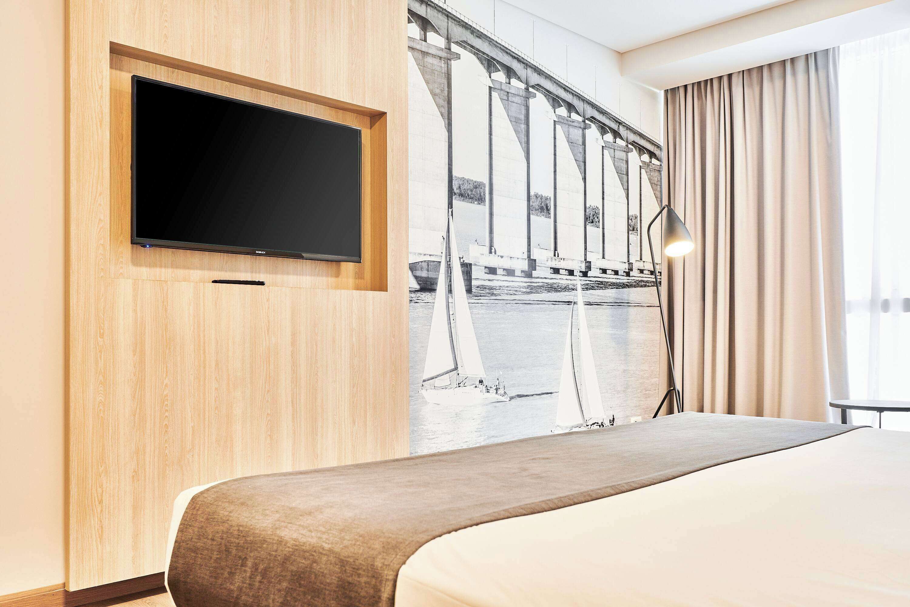 A hotel room with a large bed, a flat-screen TV mounted on a wooden panel, and a mural of a bridge and sailboats on the wall.
