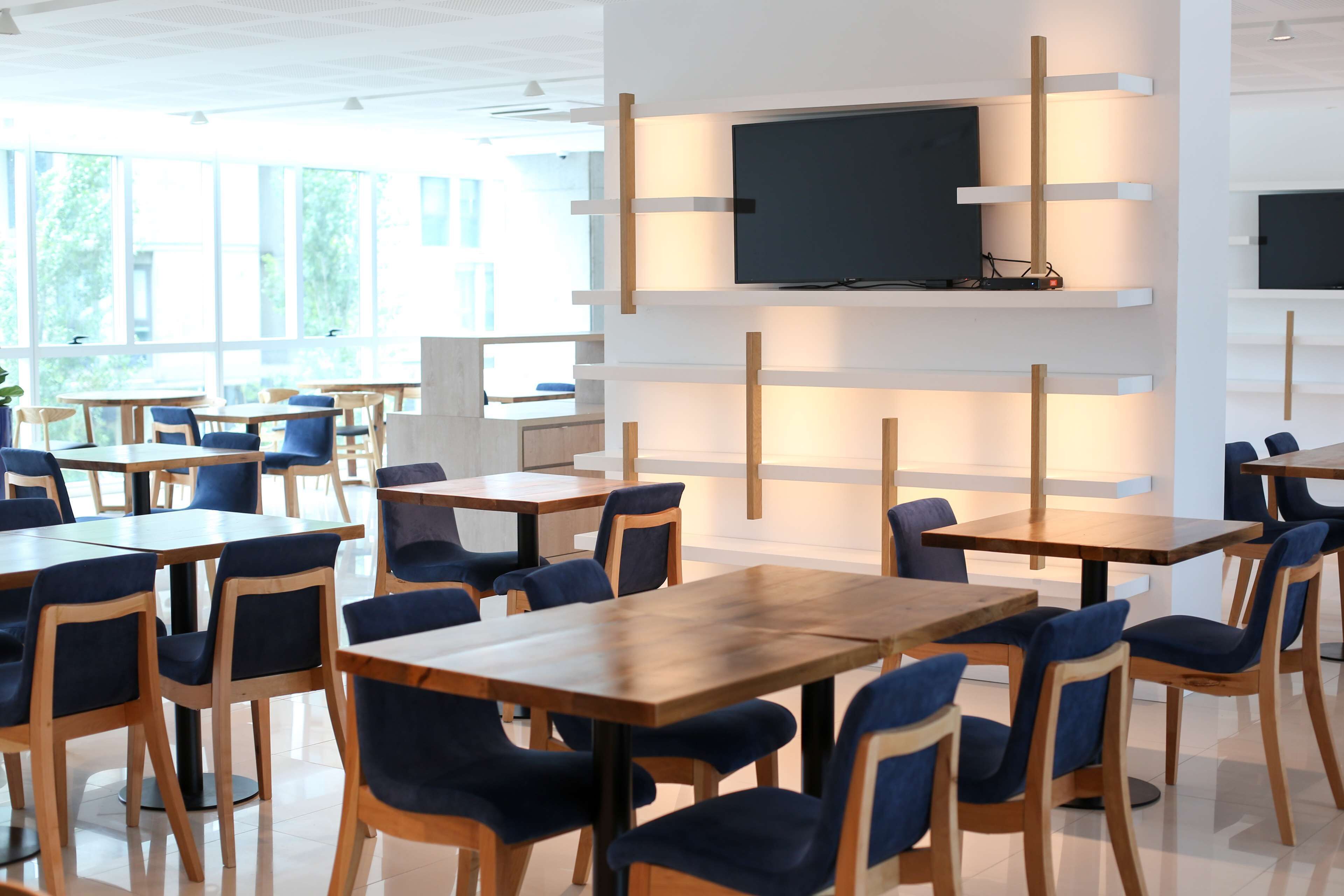 Un comedor moderno con mesas de madera y sillas tapizadas en azul, un televisor de pantalla plana montado en una pared blanca y ventanas amplias que dejan entrar luz natural.