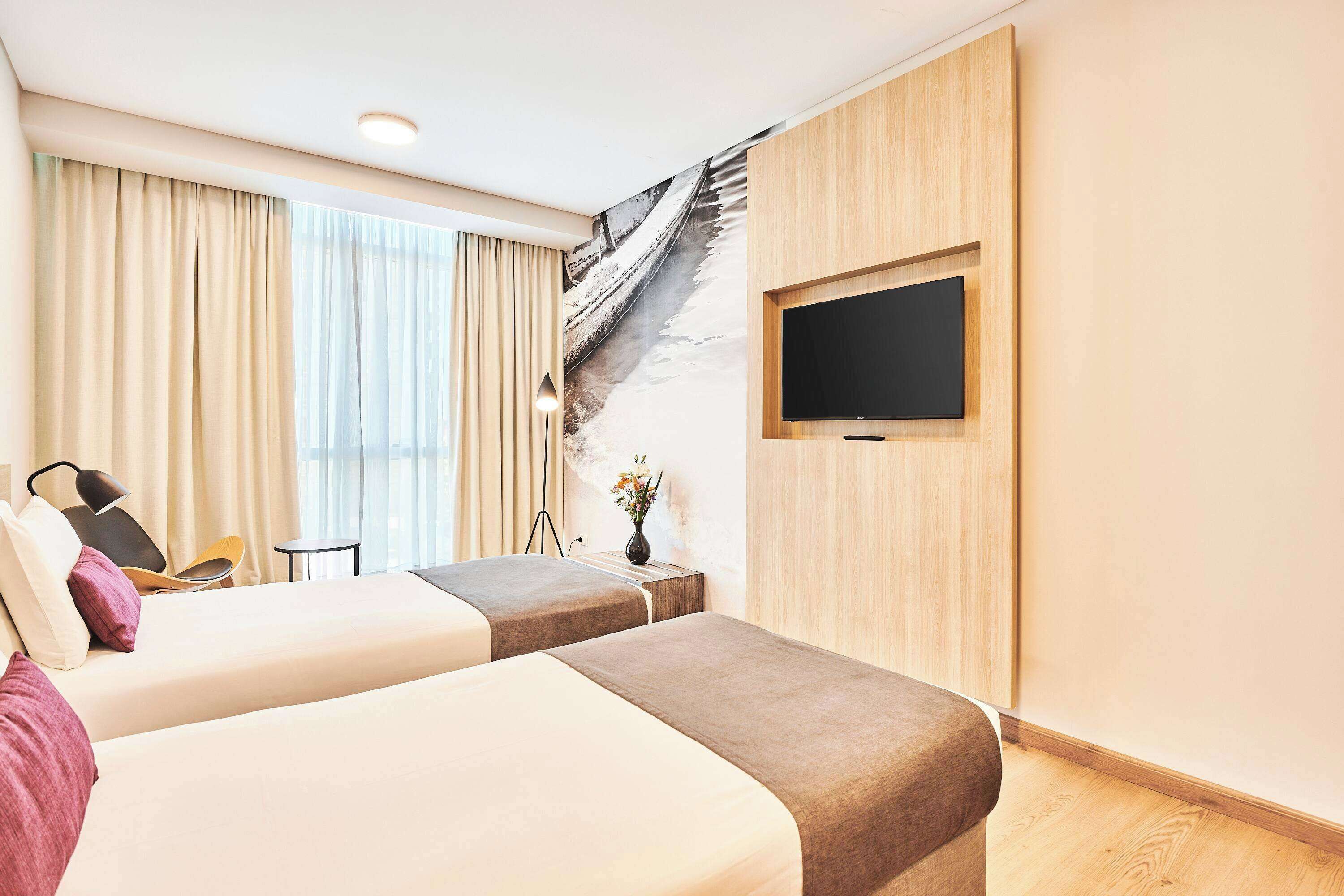 A hotel room with two beds, a TV mounted on the wall, a desk with a chair, and a large window with curtains.