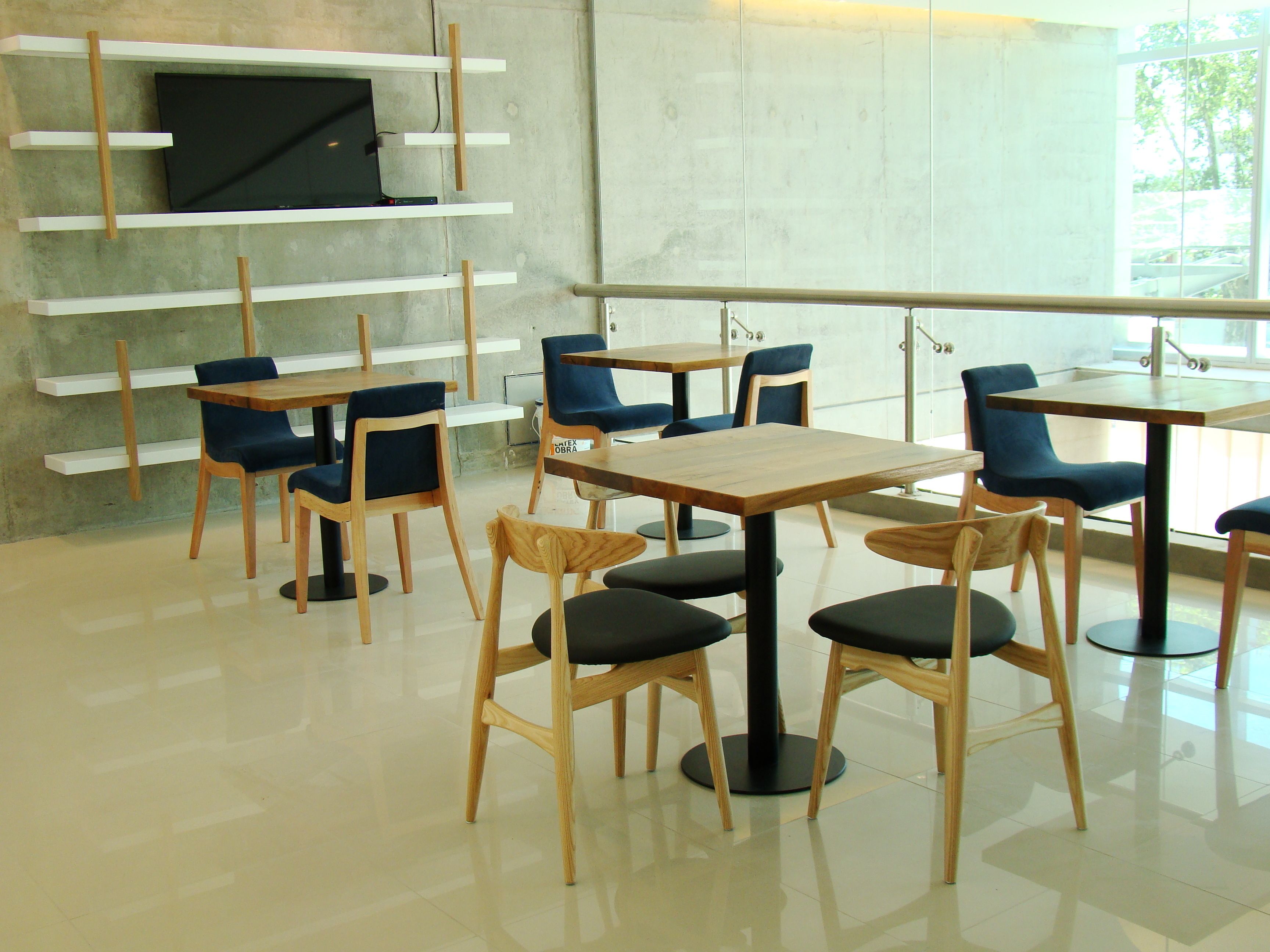 Un café moderno con mesas de madera y sillas azules, un televisor de pantalla plana sobre un estante blanco, y amplios ventanales con vistas al exterior.