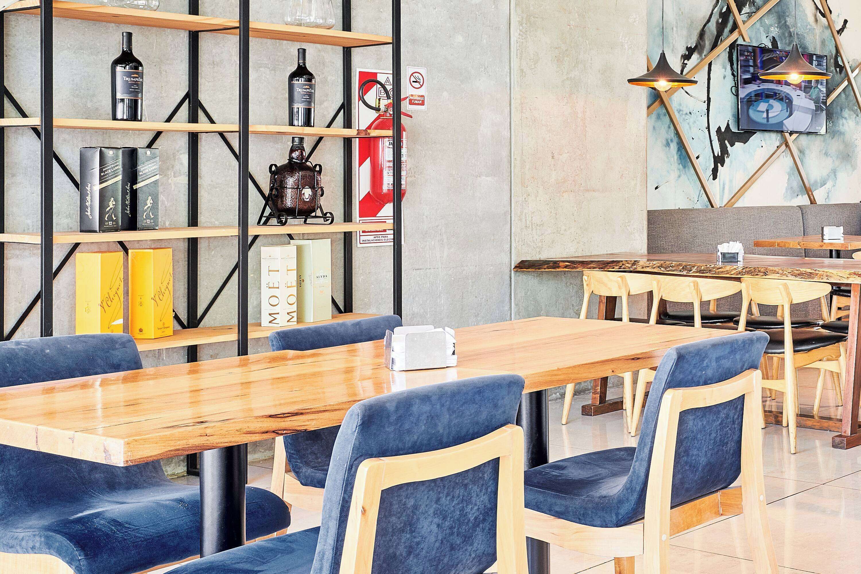 A modern cafe with wooden tables and blue upholstered chairs, a bookshelf with various items, and a wall-mounted fire extinguisher.