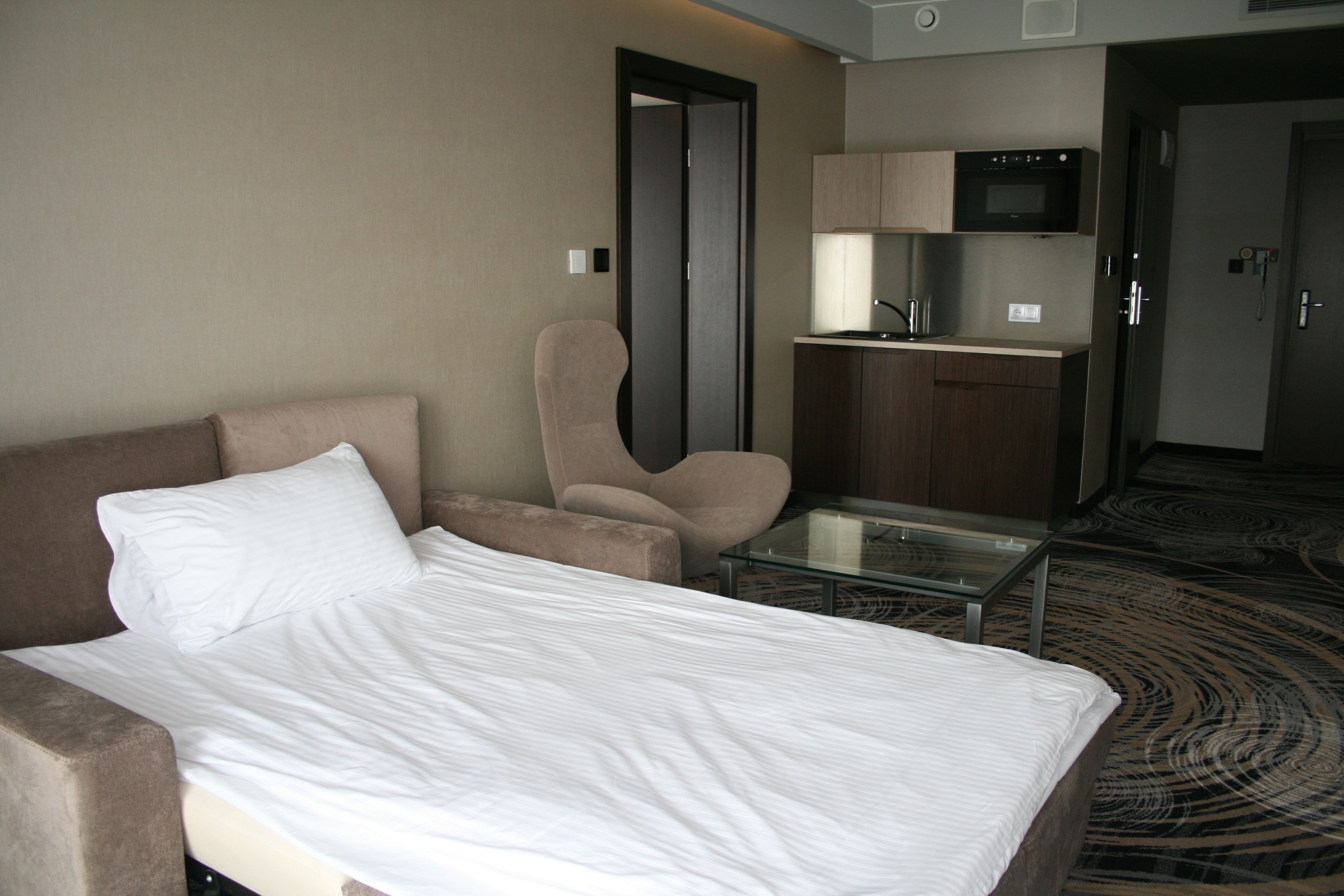 A hotel room with a bed, a chair, a glass table, and a kitchenette.