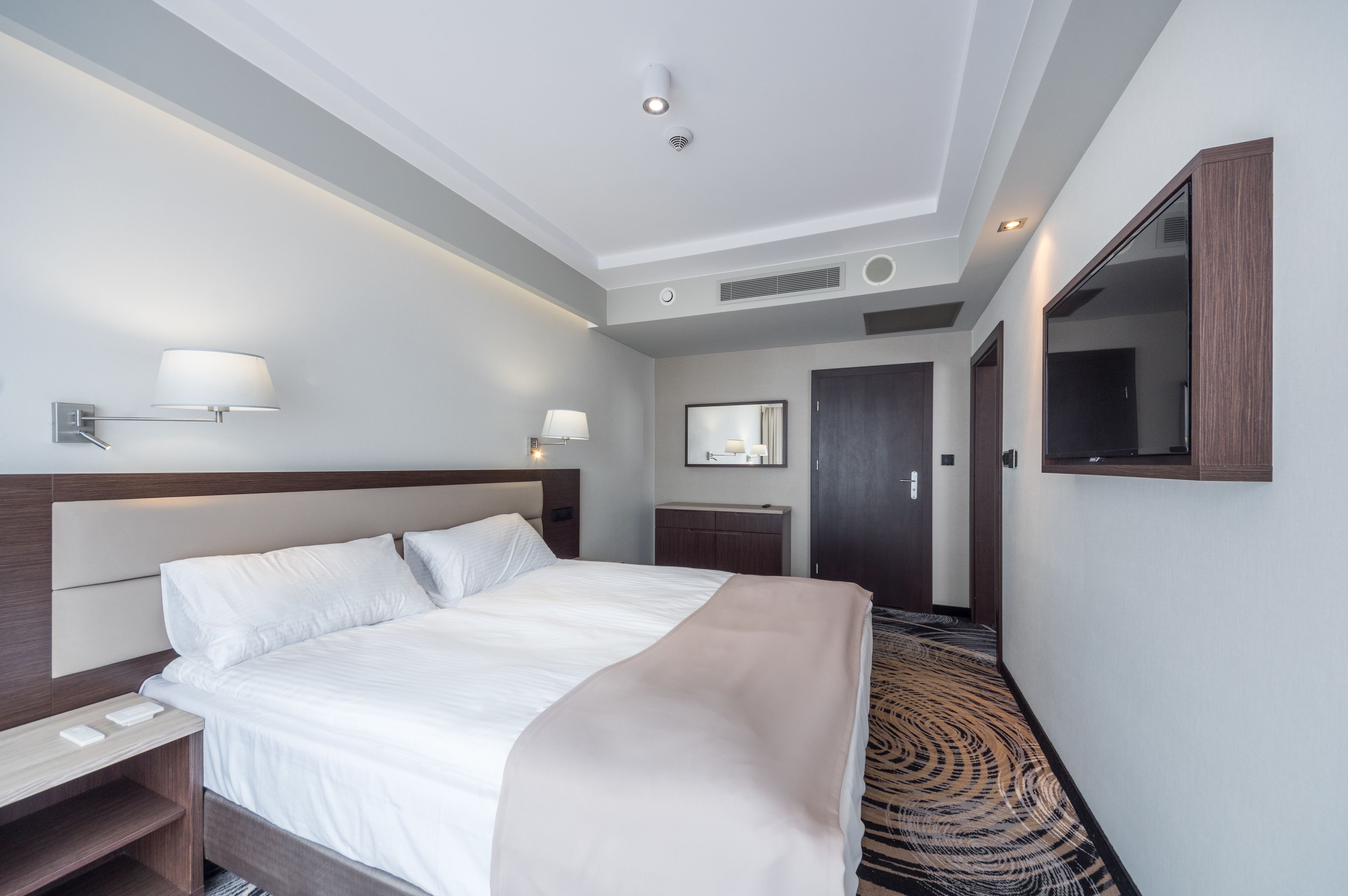A modern hotel room with a large bed, bedside tables, a desk, a mirror, and a patterned carpet.