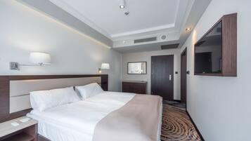 A modern hotel room with a large bed, bedside tables, a desk, a mirror, and a patterned carpet.