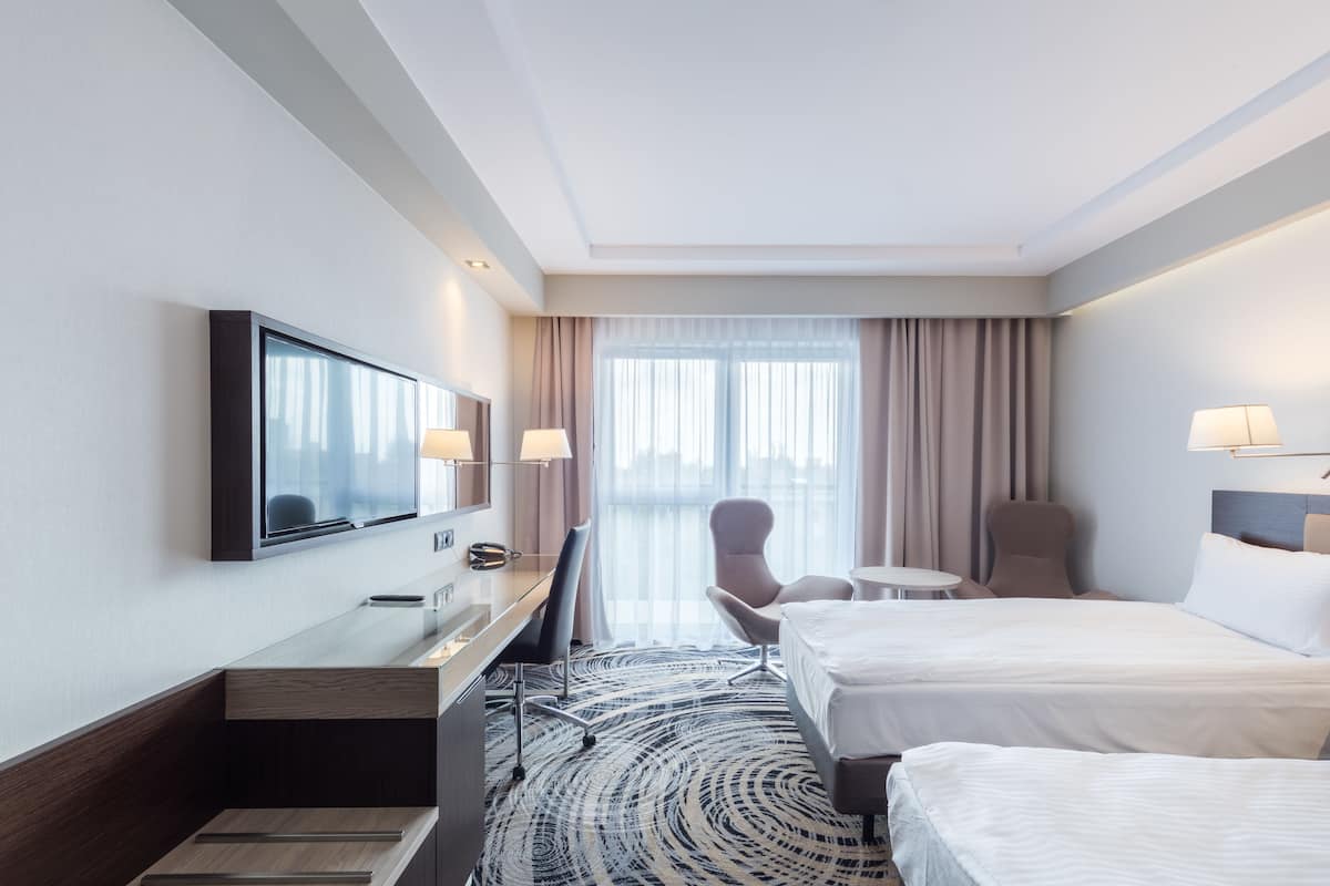 A hotel room with two beds, a desk, a chair, a TV, and a view of the city through large windows.