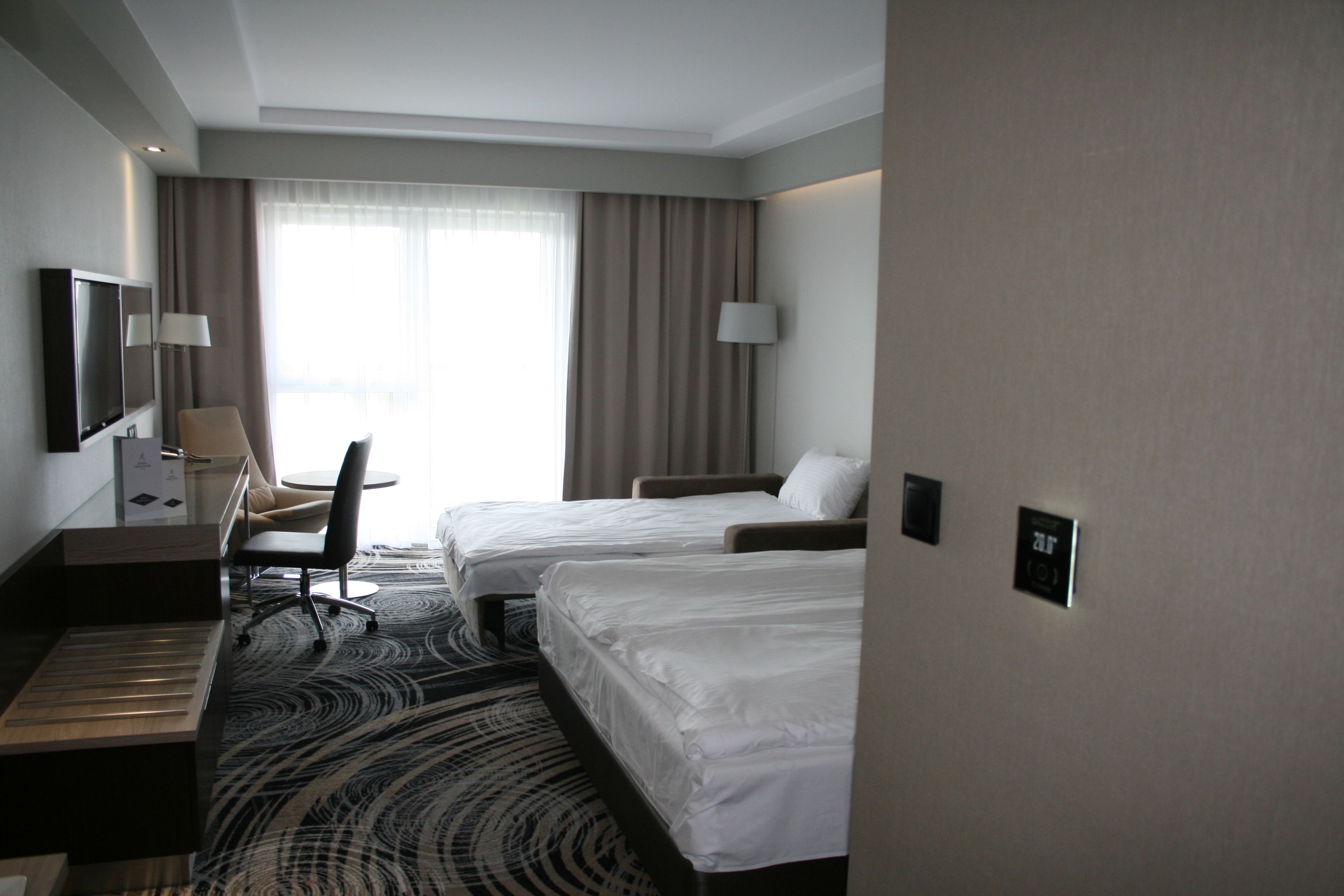A hotel room with two beds, a desk, a chair, a TV, and a large window with curtains.