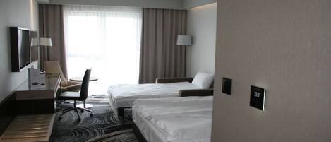 A hotel room with two beds, a desk, a chair, a TV, and a large window with curtains.