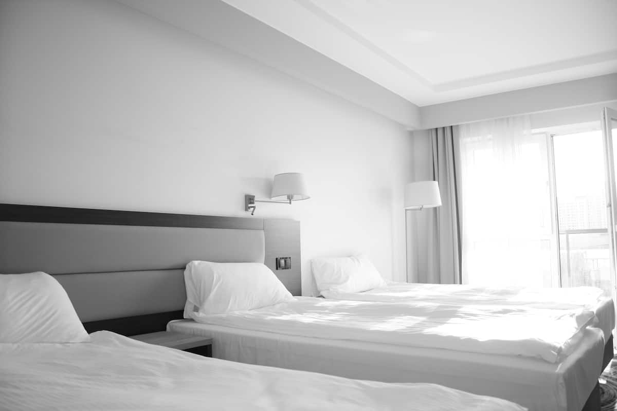 A hotel room with two beds, a nightstand, a wall-mounted lamp, and a window with curtains.