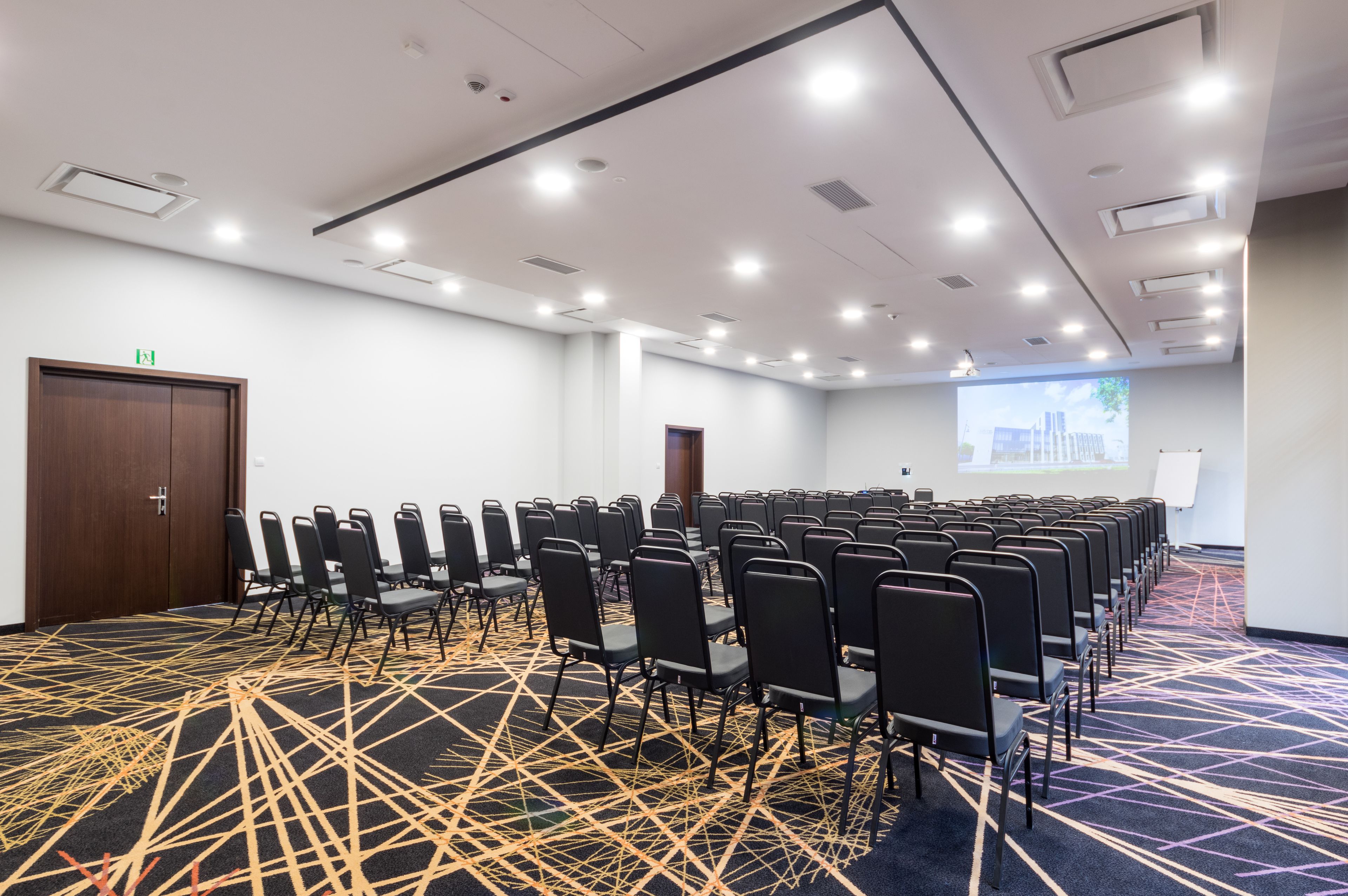 A conference room with rows of chairs, a projector screen, and a carpet with a geometric pattern.