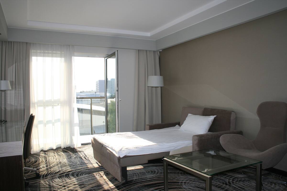 A hotel room with a bed, a glass coffee table, a chair, a desk, and a view of the city through large windows.