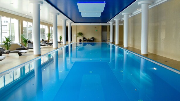 Una amplia piscina cubierta con agua cristalina y azul, rodeada de columnas blancas y tumbonas.