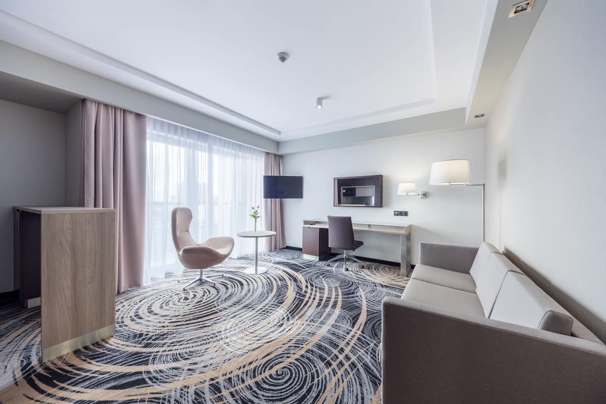 A hotel room with a large window, a desk, a flat-screen TV, a sofa, and a patterned carpet.