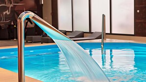 An indoor swimming pool with clear water, a metal pool ladder, lounge chairs, and a wall with abstract art.