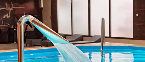 An indoor swimming pool with clear water, a metal pool ladder, lounge chairs, and a wall with abstract art.