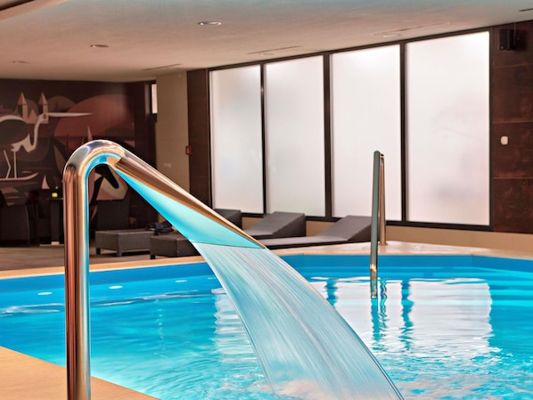 Una piscina interior con agua cristalina, una escalera metálica, sillas de descanso y una pared con arte abstracto.