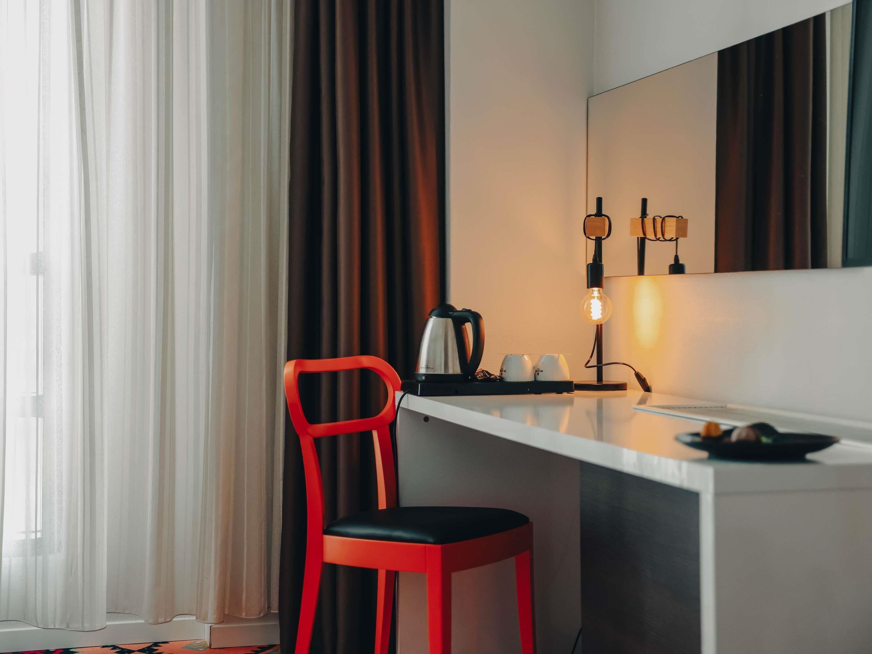 Une chambre d’hôtel avec un bureau blanc, une chaise rouge, une machine à café et une lampe.