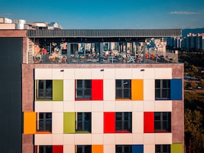 Un edificio de varios pisos con una terraza en la azotea, mobiliario exterior y vistas panorámicas de la ciudad.