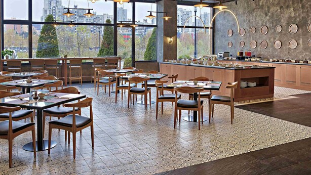 A restaurant with a high ceiling, exposed ductwork, and large windows offering a city view. Tables are arranged with chairs, and a counter with a variety of dishes is visible.
