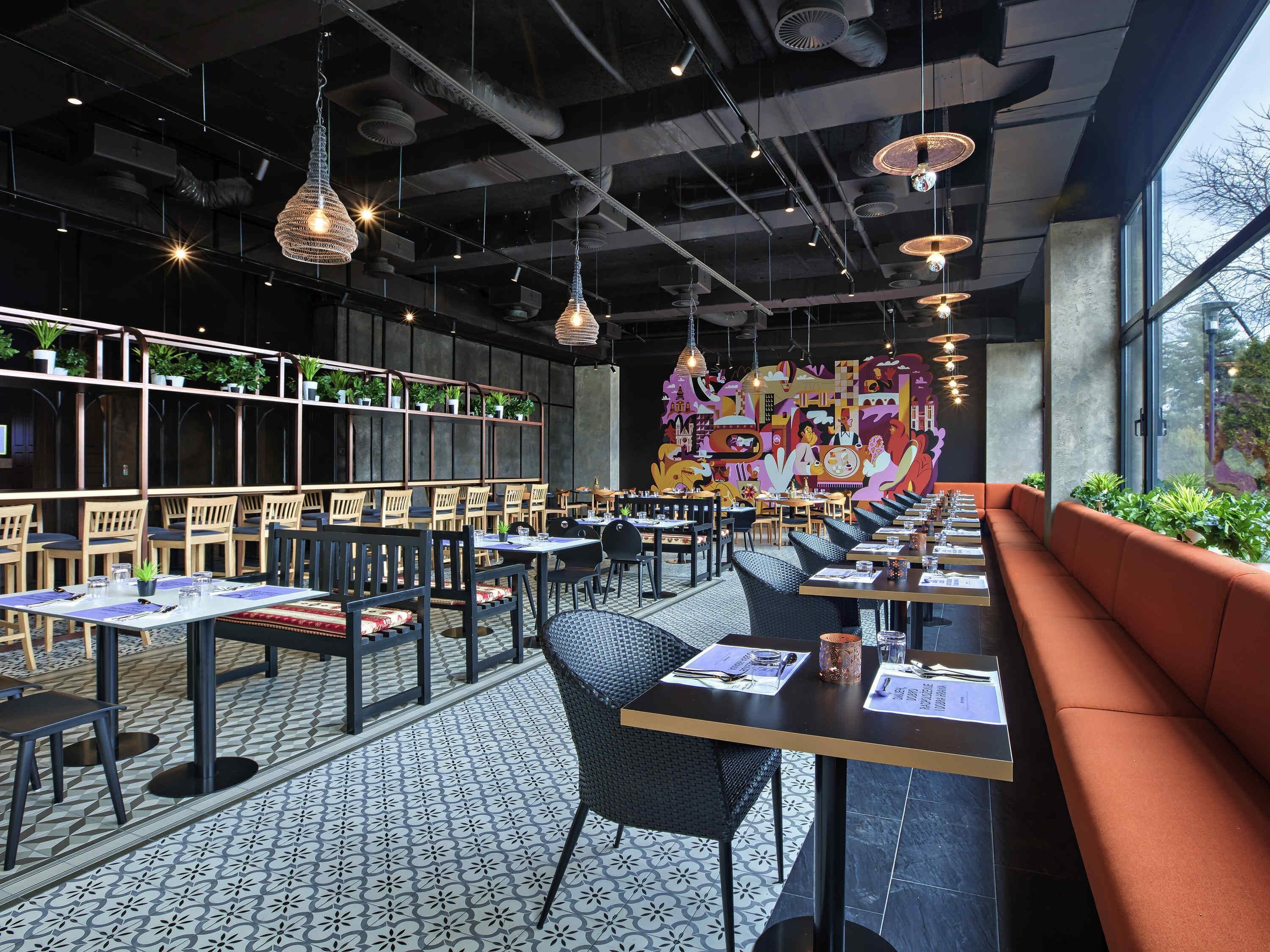 A modern restaurant interior with high ceilings, hanging pendant lights, and a colorful mural on the back wall. Tables are arranged for dining, and there are potted plants on shelves.