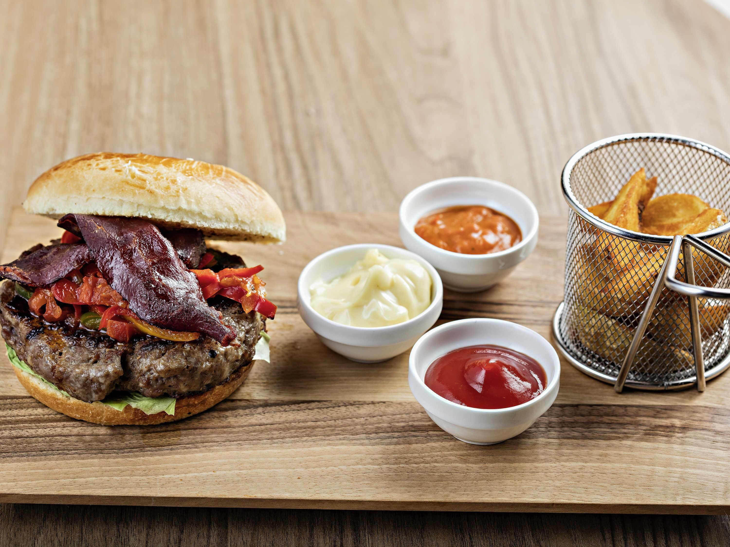 Een hamburger met spek, sla en tomaat, vergezeld van kleine schaaltjes sauzen en een mand friet.