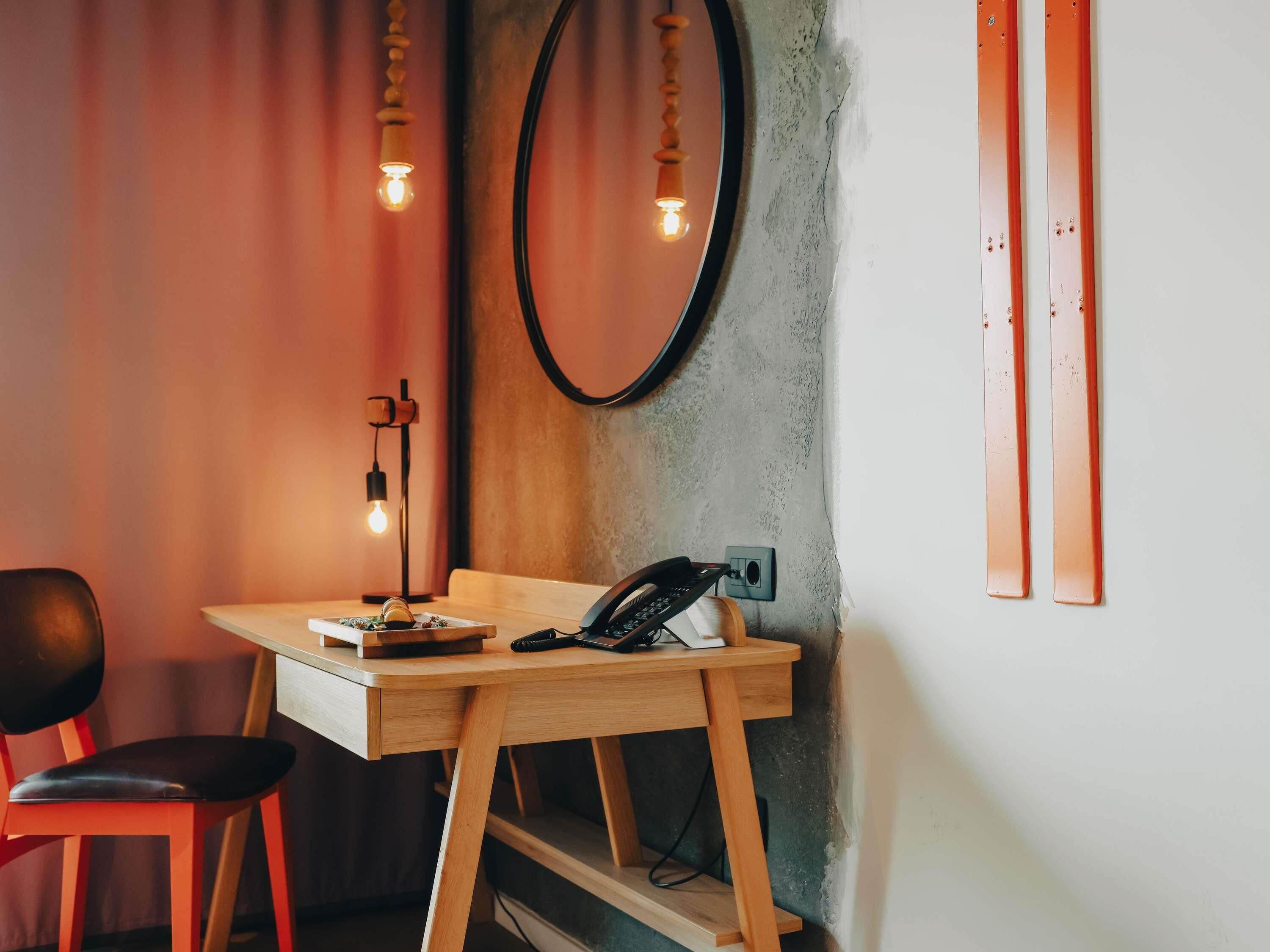 A small wooden desk with a telephone, a lamp, and a chair in a room with a concrete wall and a round mirror.