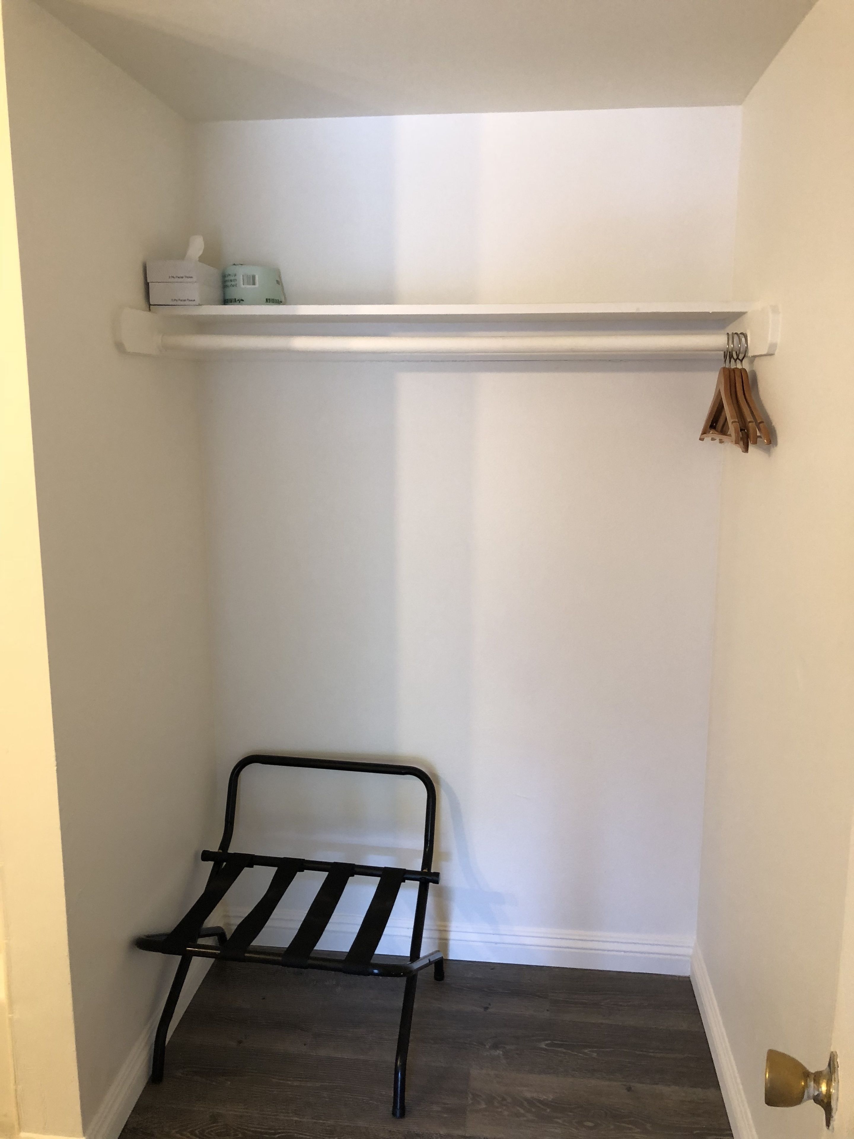 Quarto pequeno e estreito com um banco de metal preto, uma estante branca com uma caixa de lenços e uma garrafa, e uma porta com puxador de latão.